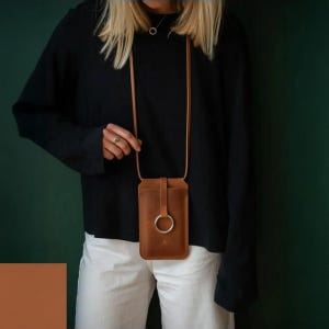 May include: A tan leather phone pouch with a long, thin shoulder strap and a prominent silver-toned metal ring detail on the front flap. The rectangular pouch features a vertical strap closure that secures through the central metal ring.