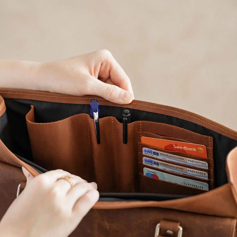 May include: A brown leather bag interior featuring a black fabric lining, two vertical pen loops, and a series of horizontal card slots containing three credit cards, including one clearly labeled with the text Swedbank.