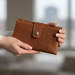 以下が含まれることがあります： スナップ留めと刻印されたイニシャルが付いた茶色の革製財布。長方形の財布には、シルバーのスナップで固定されたフラップが付いています。「M.A.L」のイニシャルが刻印されています。