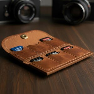 Puede incluir: Un tarjetero de cuero marrón para tarjetas SD con múltiples ranuras. El tarjetero está abierto, revelando varias tarjetas SD de varios colores. Un cierre de botón de latón asegura el tarjetero. Dos cámaras están borrosas en el fondo.