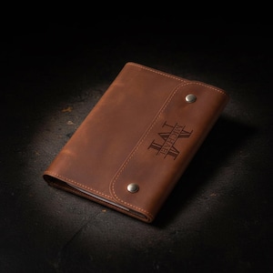 Puede incluir: Una funda de cuero marrón para cuaderno con cierre de solapa asegurado con dos broches de metal plateado. La parte frontal de la funda tiene grabado el nombre MICHAEL y la letra M en una fuente estilizada. El cuero tiene un acabado mate.