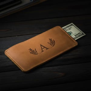 Può includere: Un portafoglio in pelle marrone chiaro con la lettera "A" e un disegno di grano inciso sul davanti. Una banconota da cento dollari è parzialmente visibile all'interno. Il portafoglio è rettangolare e ha un bordo cucito.