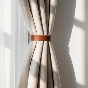 May include: A pair of beige linen curtains held together by a brown leather curtain tieback. The tieback is a simple band that wraps around the curtains, creating a neat and organized look. The curtains are hanging in front of a white wall.