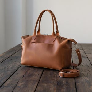 May include: A tan leather tote bag featuring two top handles, a zippered main compartment, a horizontal slit pocket on the front, and side pockets. A detachable, adjustable shoulder strap with silver-toned metal hardware lies beside the bag.