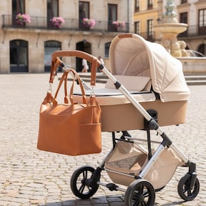May include: A beige baby stroller with a silver frame and a large, tan leather-like diaper bag hanging from the handle. The stroller features a protective canopy, a bassinet-style seat, four black wheels, and a storage basket underneath the frame.