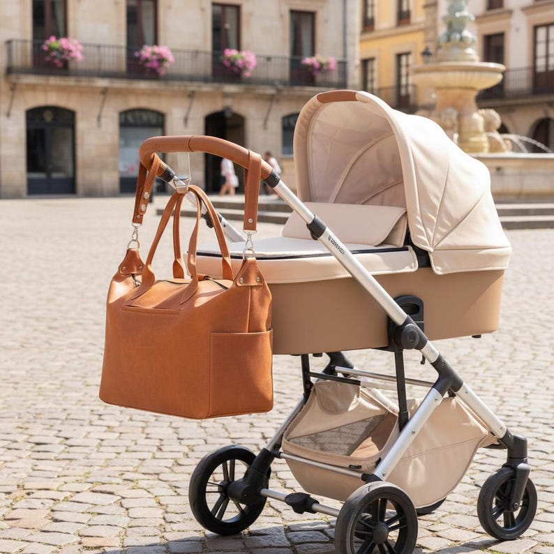 May include: A beige baby stroller with a silver frame and a large, tan leather-like diaper bag hanging from the handle. The stroller features a protective canopy, a bassinet-style seat, four black wheels, and a storage basket underneath the frame.