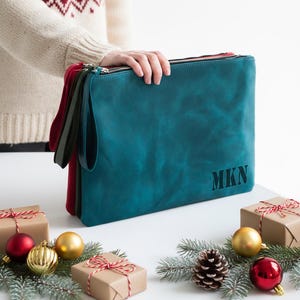 May include: Teal leather clutch bag with the initials "MKN" embossed on the front. The clutch has a wrist strap and a zip closure. A red and green clutch are visible behind the teal one. Christmas decorations and wrapped gifts are in the foreground.
