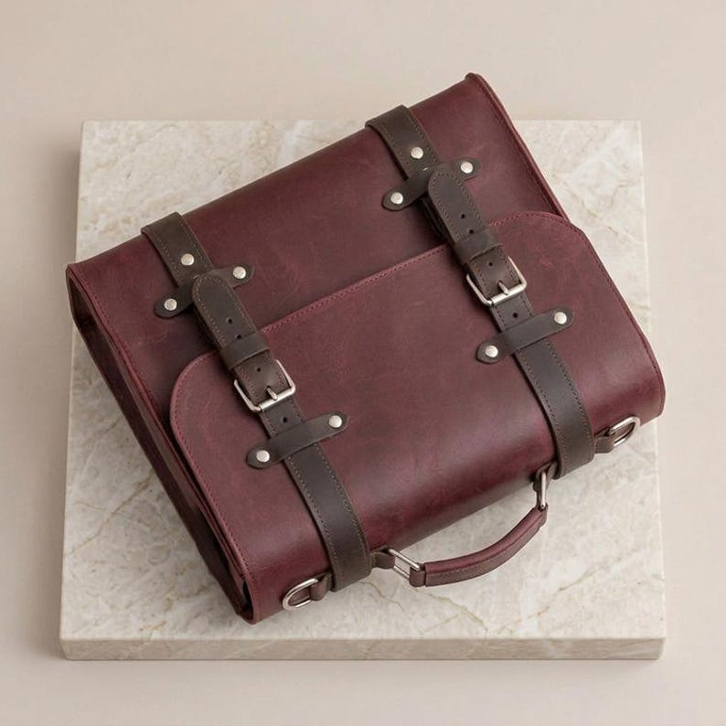 May include: A deep burgundy leather satchel featuring two dark brown leather straps with silver-toned metal buckles and rivets. The bag includes a top handle, a front flap closure, and side metal D-rings for attaching a shoulder strap.