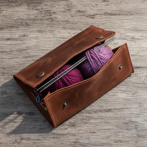 May include: A brown leather case with a flap secured by two brass-colored metal snaps. Inside the case are two balls of purple yarn, several long silver knitting needles, and a small collection of colorful plastic stitch markers for knitting.