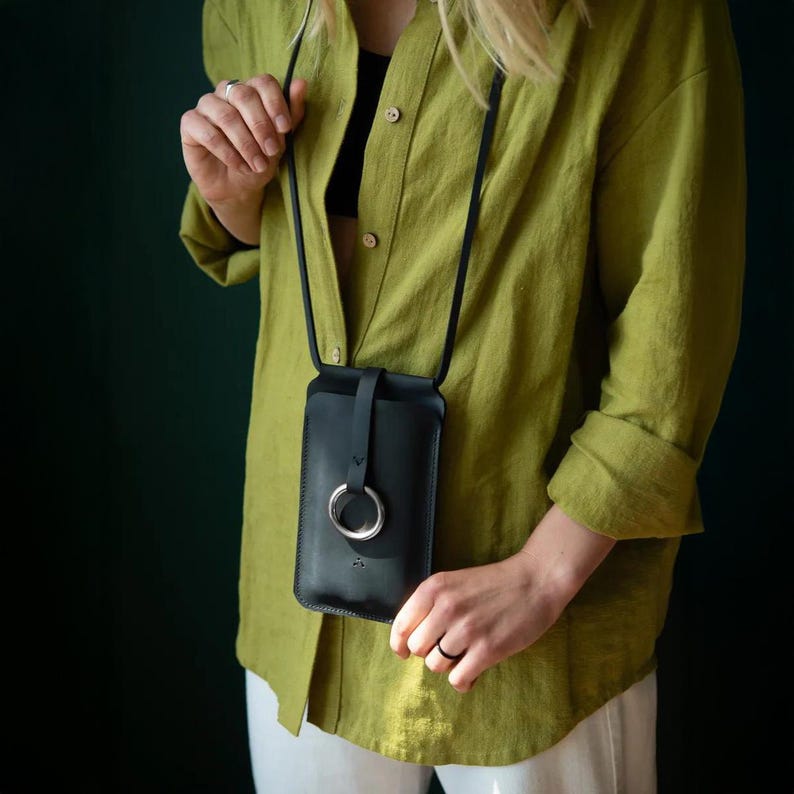 May include: A black leather phone pouch with a long thin strap and a silver metal ring closure on the front. The pouch is worn over a lime green button-down shirt and white trousers, featuring a smooth finish and a minimalist vertical design.