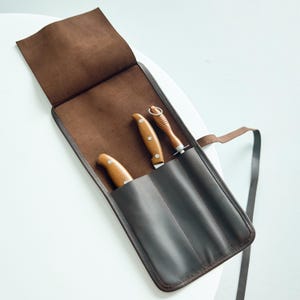 Op de afbeelding: Een bruine leren messenrol met drie messen. De messen hebben houten handvatten en roestvrijstalen lemmeten. De rol is open en toont de messen in afzonderlijke vakken. Het leer is een diepe, rijke bruine kleur.