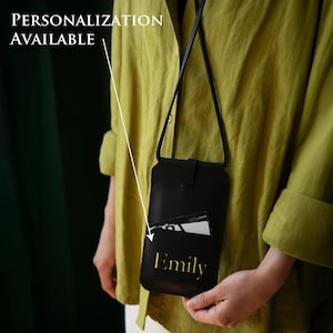 May include: Black leather crossbody phone bag with a front pocket holding a card. The bag has a long strap and a flap closure. The name "Emily" is printed in gold on the front. Text on the image reads "Personalization Available".