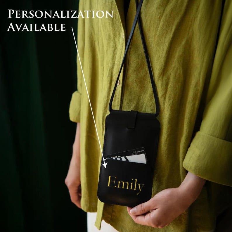 May include: Black leather crossbody phone bag with a front pocket holding a card. The bag has a long strap and a flap closure. The name "Emily" is printed in gold on the front. Text on the image reads "Personalization Available".