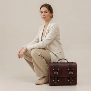 May include: A dark burgundy leather satchel featuring two vertical dark brown straps with silver-toned metal buckles and rivets. The bag has a top handle and a rectangular flap closure, showcasing a structured design with visible stitching details.