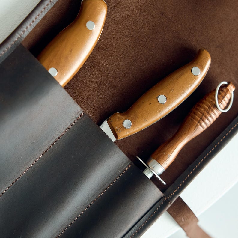 May include: Close-up of a brown leather knife roll with three knives. The knives have wooden handles and silver rivets. The roll is dark brown and the knives are partially visible.