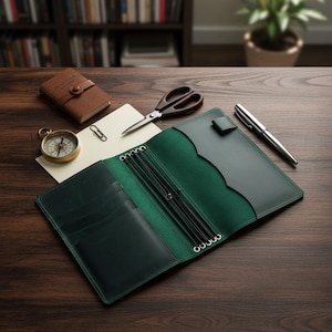 Puede incluir: Un diario de cuero verde oscuro con anillos plateados y un bolígrafo a juego descansa sobre una superficie de madera. Un pequeño cuaderno marrón, tijeras, una brújula y un clip sugieren un tema de papelería.