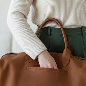 May include: A large, smooth, tan leather duffel bag featuring two sturdy rounded handles and a horizontal zippered pocket on the front exterior. The bag is constructed with clean lines and a minimalist aesthetic, showcasing a rich, warm brown tone.