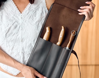 May include: A dark brown leather knife roll featuring three vertical slots. Two slots contain knives with light brown wooden handles, while the third slot holds a tool with a ribbed wooden handle and a small metal ring at the top end.