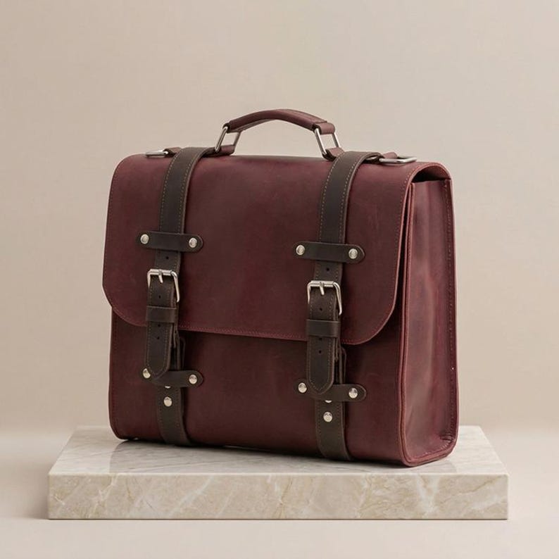 May include: A deep burgundy leather satchel featuring a top handle and two vertical dark brown leather straps with silver-toned metal buckles and rivets. The bag has a structured rectangular shape with a flap closure and visible stitching.