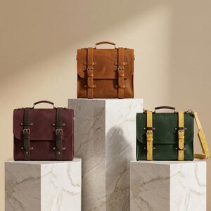 May include: Three rectangular satchel bags with top handles and dual buckle straps. One bag is tan with matching straps, one is deep burgundy with dark brown straps, and one is forest green with bright yellow straps, each featuring metal hardware.
