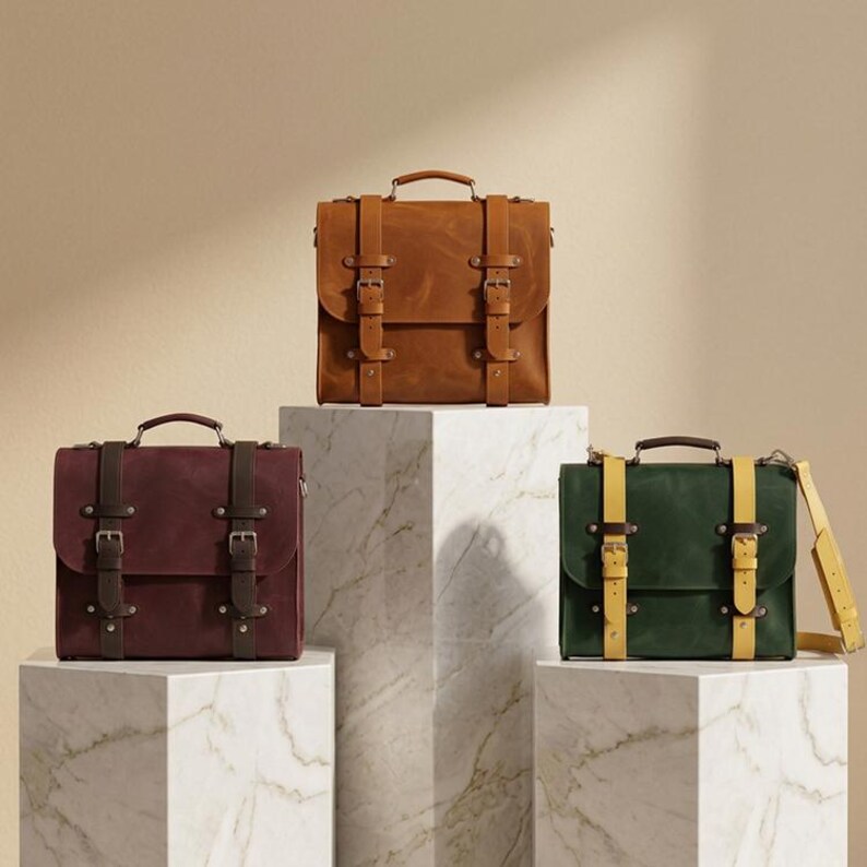 May include: Three rectangular satchel bags with top handles and dual buckle straps. One bag is tan with matching straps, one is deep burgundy with dark brown straps, and one is forest green with bright yellow straps, each featuring metal hardware.