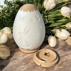 Può includere: Un oggetto decorativo a forma di uovo, bianco sporco, con base e parte superiore beige. L'uovo presenta disegni in rilievo di farfalle, fiori e un coniglietto. Il coperchio è staccato. Posizionato su una superficie di legno.