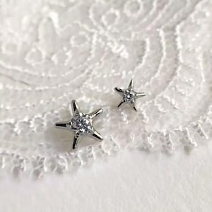 May include: Two silver star-shaped stud earrings with a clear, sparkling center stone. The earrings are set against a white, textured background, creating a contrast that highlights the jewelry's design. These earrings are a delicate accessory.