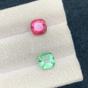 May include: Two cushion-cut gemstones. One is a vibrant pink-red color, and the other is a light green. Both are set on a white surface, with a dark background. The gems are likely for jewelry making or collecting.