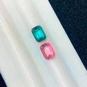 Può includere: Due pietre preziose rettangolari. Una è di un verde-azzurro vibrante, l'altra rosa tenue. Entrambe le pietre sono poste su una superficie bianca, con uno sfondo blu scuro. Le gemme sono probabilmente per la creazione di gioielli o da collezione.