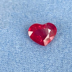 以下が含まれることがあります： 鮮やかな赤色でファセットカットが施されたハート型の宝石。尖った底部と中央がくぼんだ上部を持ち、複数の幾何学的なカットが光を反射して、磨き上げられた半透明の赤い表面を際立たせています。