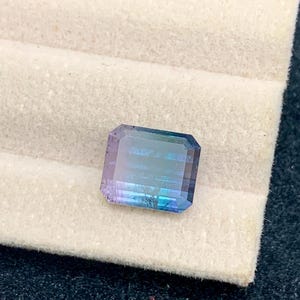 May include: A rectangular gemstone with a gradient of colors, including blue, purple, and green. The gem is set against a white, textured background. The gemstone is a beautiful example of natural mineral beauty.