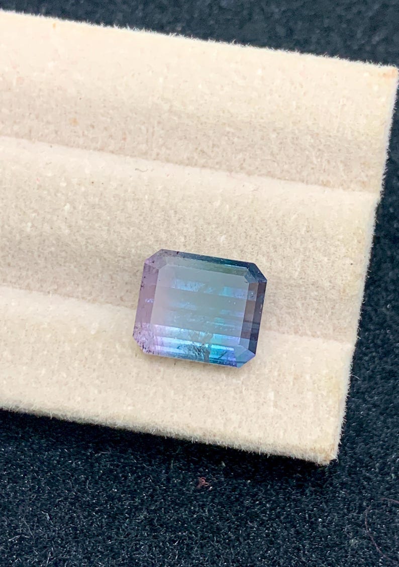 May include: A rectangular gemstone with a gradient of colors, including blue, purple, and green. The gem is set against a white, textured background. The gemstone is a beautiful example of natural mineral beauty.