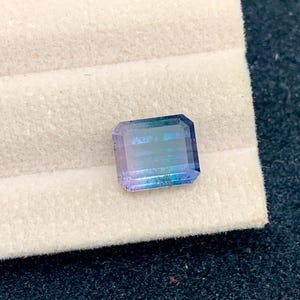 May include: A rectangular gemstone with a gradient of colors, transitioning from purple to blue and green. The gem is set against a white, textured background, showcasing its facets and clarity. The stone is likely a tourmaline.