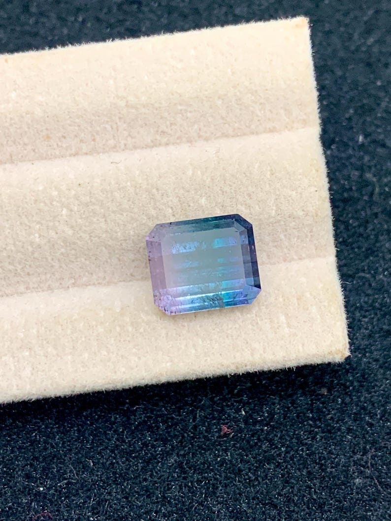 May include: A rectangular gemstone with a gradient of colors, transitioning from purple to blue and green. The gem is set against a white, textured background, showcasing its facets and clarity. The stone is likely a tourmaline.