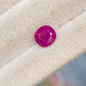 May include: A faceted, oval ruby gemstone. The gem is a deep, vibrant red colour, with multiple facets catching the light. The ruby is set against a light-coloured, textured background.