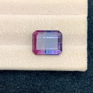 May include: An emerald-cut gemstone with a gradient of colors, transitioning from deep pink to purple and blue. The gem is set against a white background, showcasing its facets and clarity. This gemstone is likely for jewelry.
