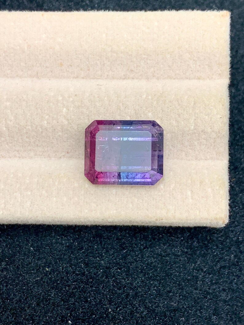 May include: An emerald-cut gemstone with a gradient of colors, transitioning from deep pink to purple and blue. The gem is set against a white background, showcasing its facets and clarity. This gemstone is likely for jewelry.