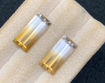 May include: Two rectangular gemstones with a gradient of colors, transitioning from a golden yellow to a clear, almost white hue. The gems are set on a light beige fabric, with a dark background. The gems are likely for jewelry.