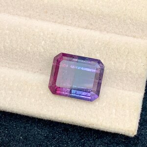May include: A rectangular gemstone with a gradient of colors, transitioning from pink to purple to blue. The gem is set against a white fabric, with a dark blue textured background.