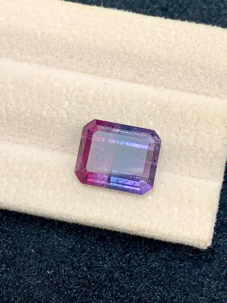 May include: A rectangular gemstone with a gradient of colors, transitioning from pink to purple to blue. The gem is set against a white fabric, with a dark blue textured background.