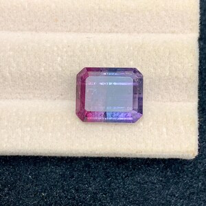 May include: A rectangular gemstone with a gradient of colors, including pink, purple, and blue. The gem is set against a cream-colored surface, showcasing its facets and clarity. The gem is a vibrant and eye-catching piece.