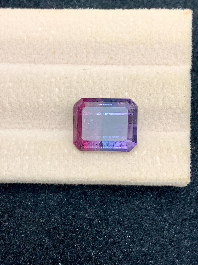 May include: A rectangular gemstone with a gradient of colors, including pink, purple, and blue. The gem is set against a cream-colored surface, showcasing its facets and clarity. The gem is a vibrant and eye-catching piece.