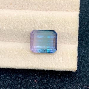 May include: A rectangular gemstone with a gradient of colors, including blue, purple, and green. The gem is faceted, with visible lines across its surface. It sits on a light beige surface, with a dark background.