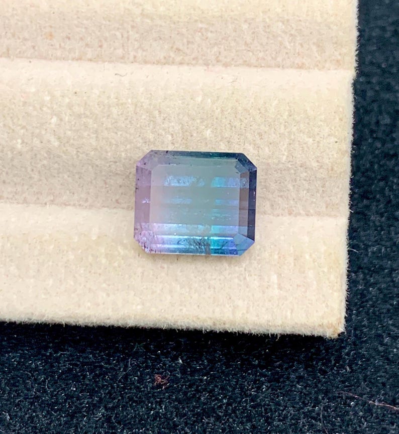 May include: A rectangular gemstone with a gradient of colors, including blue, purple, and green. The gem is faceted, with visible lines across its surface. It sits on a light beige surface, with a dark background.