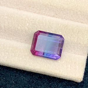 May include: A rectangular-cut gemstone with a gradient of colors, transitioning from pink to purple to blue. The gem is set on a cream-colored fabric, with a dark blue background.
