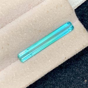 May include: A rectangular, faceted gemstone with a gradient of blue and green hues. The gemstone is displayed on a beige fabric, with a white ribbon visible in the background. The gem's shape is elongated, with visible facets.