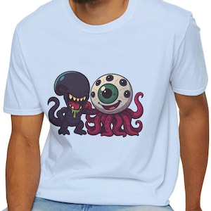 May include: Light blue t-shirt featuring a cartoon graphic of a purple alien and an eyeball-octopus hybrid. The alien has sharp teeth and the eyeball has multiple pupils. The shirt is made of a soft material.