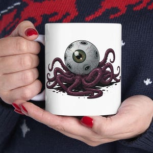 May include: White ceramic mug featuring a detailed illustration of a large eyeball with multiple smaller eyes and dark purple tentacles. The design is set against a clean white background, creating a striking contrast.