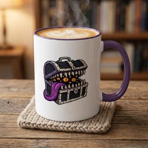 May include: White ceramic mug with a purple handle and rim, featuring a cartoon illustration of a treasure chest with multiple eyes and a long purple tongue. The mug contains a latte with latte art.