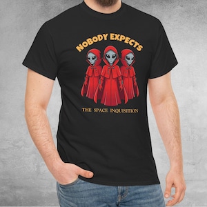 Puede incluir: Camiseta negra con un diseño de tres extraterrestres con capas rojas. El texto "NOBODY EXPECTS" está sobre los extraterrestres, con "THE SPACE INQUISITION" debajo. El diseño está centrado en la camiseta.